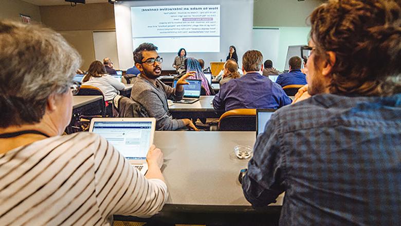 A group of people seated with laptops looking  at two presenters standing in the front of the room with an overhead presentation on the screen